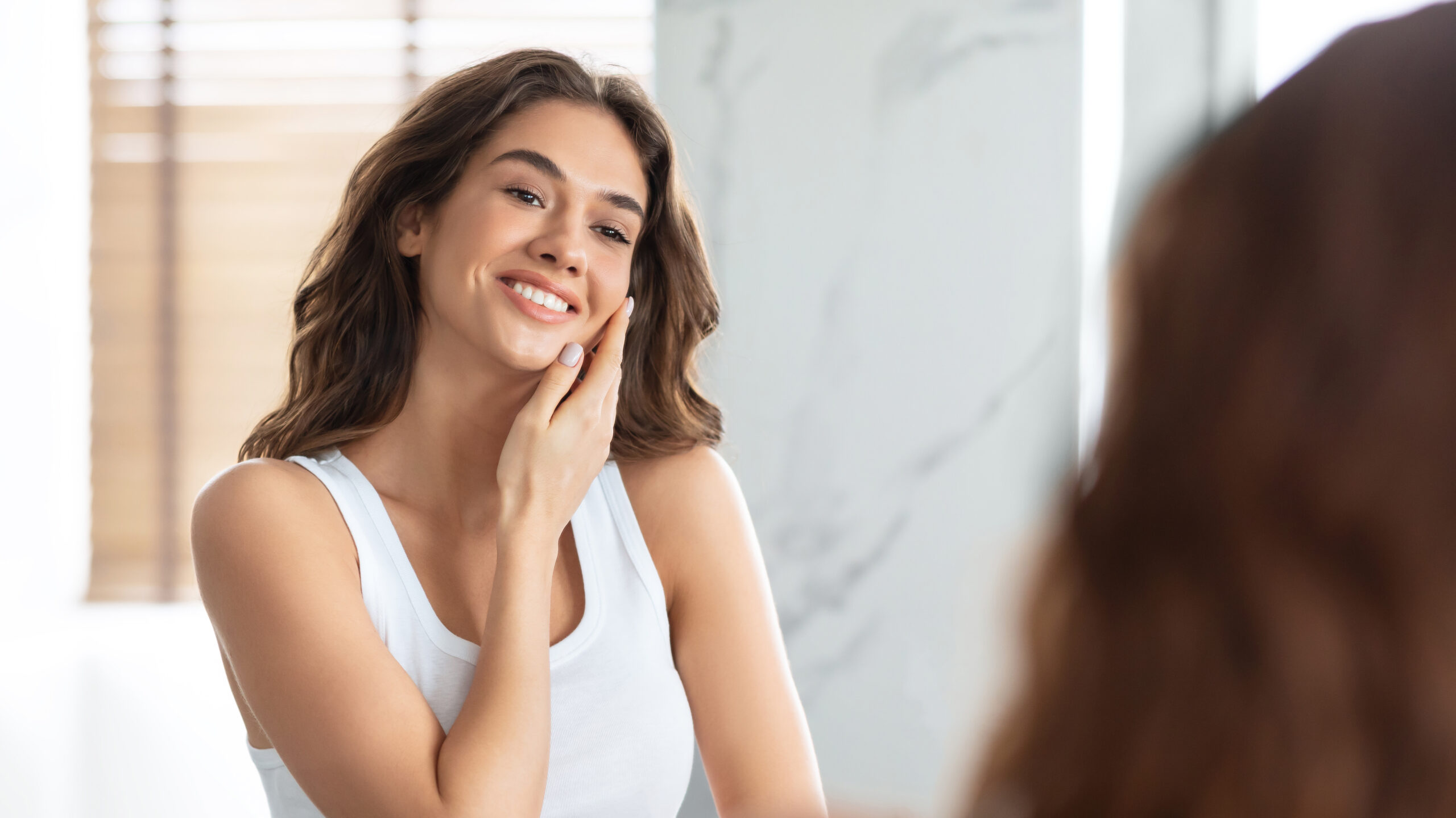 Gorgeous Young Lady Touching Face Caring For Facial Skin Standing Near Mirror In Modern Bathroom At Home. Beauty, Skincare Concept. Selective Focus, Panorama