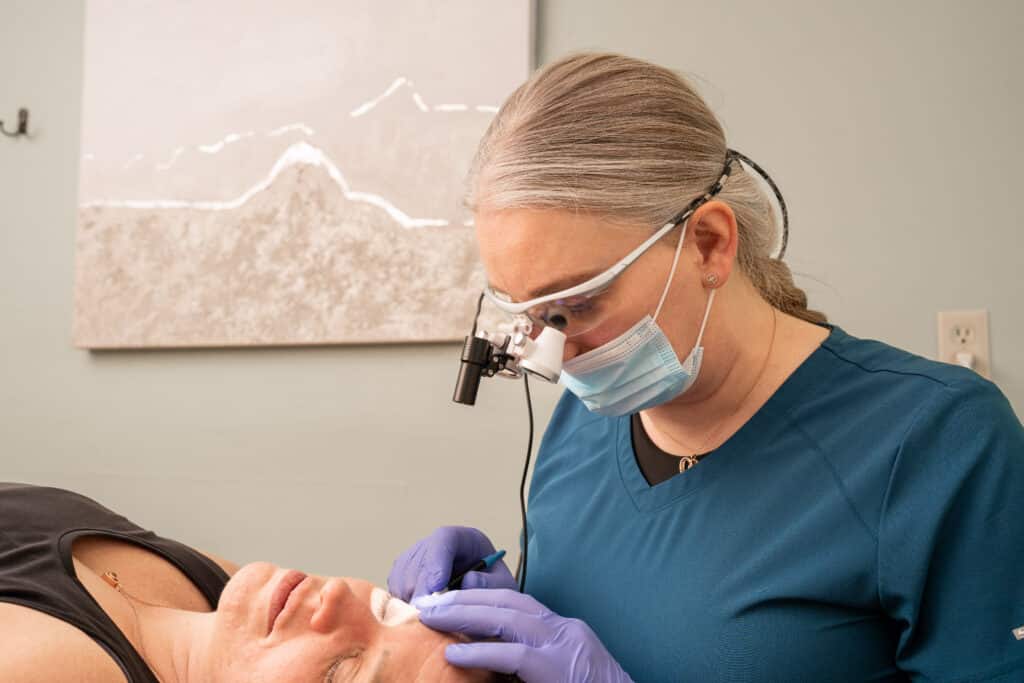 Dental professional in blue scrubs and a mask performs a procedure on a patient lying in a chair, using magnification loupes and dental tools.