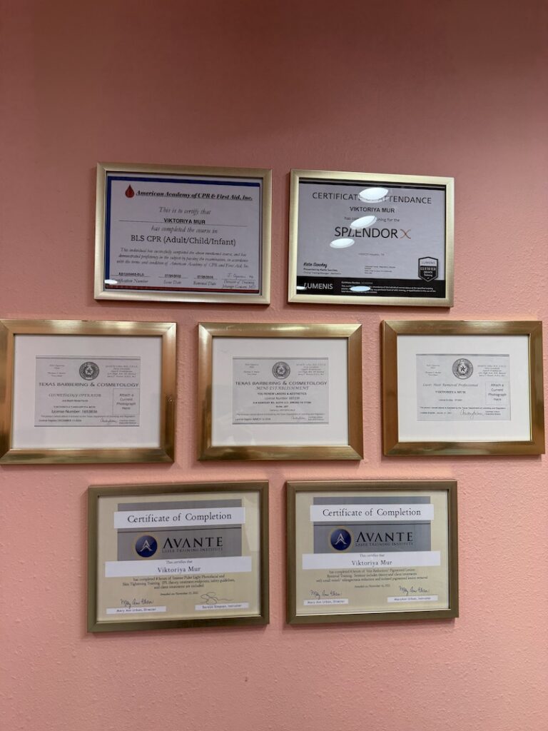Wall featuring eight framed certificates on a peach-colored wall, arranged in two rows (top row two frames, bottom row six). The frames display professional licenses and completion certificates from organizations like Avante and Texas Barbering & Cosmetology.