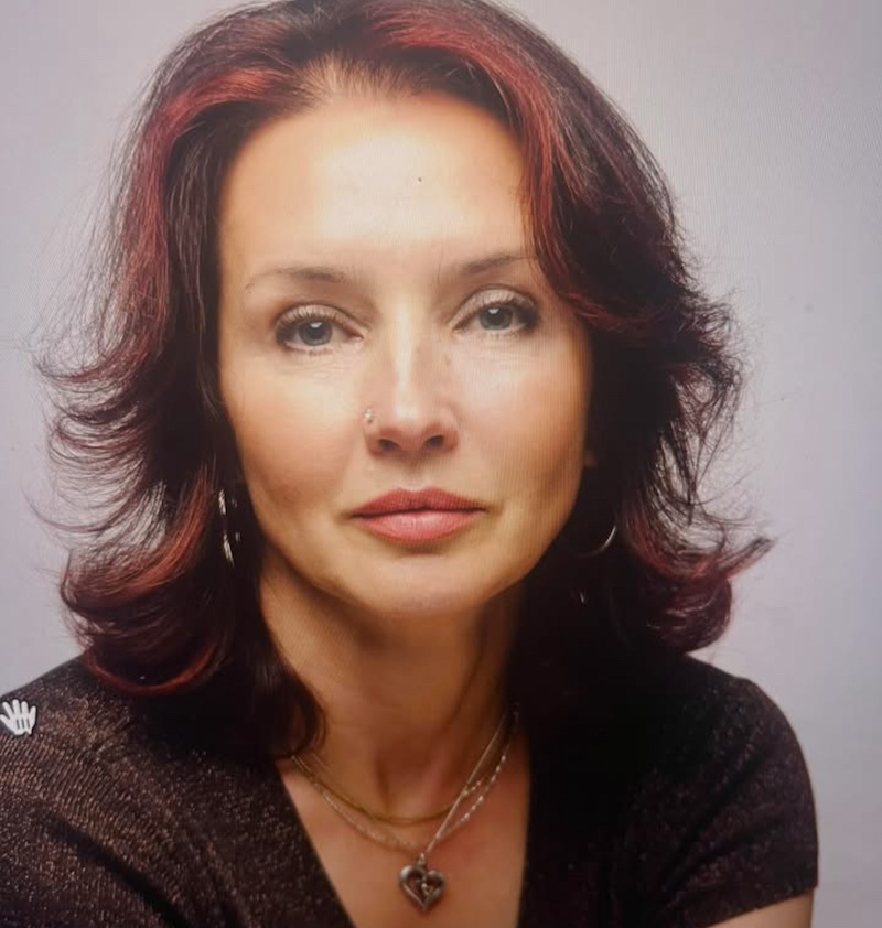 Portrait of a woman with burgundy and dark hair, wearing hoop earrings and a heart pendant necklace, looking at the camera.