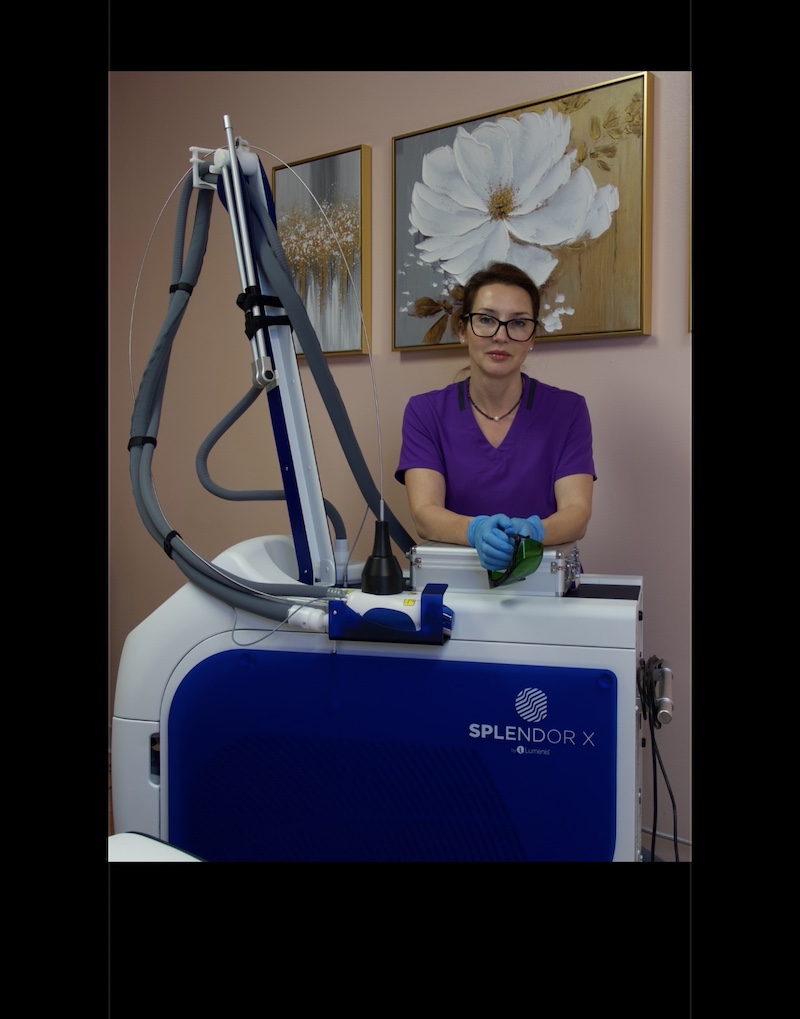 Female clinician in purple scrubs wearing blue gloves operates a SPLENDOR X laser machine in a clinic treatment room.